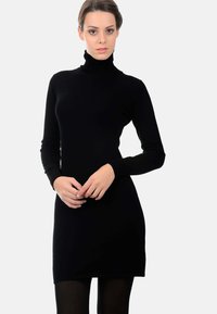 Eine Frau trägt ein enganliegendes schwarzes Rollkragenkleid mit langen Ärmeln und steht vor einem schlichten weißen Hintergrund.