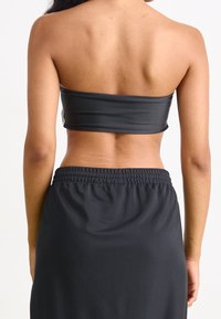 Černý top bez ramínek s hladkou, pružnou texturou. Spárovaný s černou sukní s elastickým pasem. Zobrazen pohled zezadu.