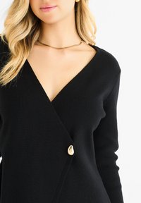 Black knitted wrap cardigan featuring a deep V-neck and a prominent gold-tone button at the waist. Soft texture and fitted sleeves.