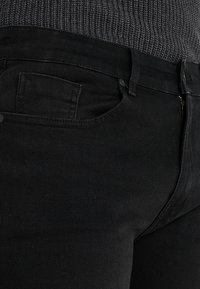 Černé džínové kalhoty s hladkou texturou, vybavené přední kapsou, zapínáním na knoflík a zipem. Pas sedí nad šedým pleteným topem.