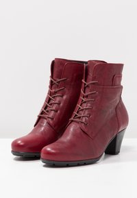 Botas de tobillo de cuero rojo con un acabado texturizado, que cuentan con cordones marrones, una puntera redondeada y un resistente tacón negro.