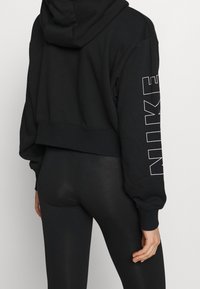 Sudadera negra recortada con capucha, que presenta letras blancas de "NIKE" en la manga izquierda. Combinada con leggings negros brillantes, que muestran una textura suave.