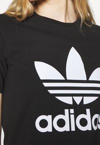 Svart bomullströja med en stor vit Adidas trefoil-logotyp som har tre bladliknande former och tre horisontella ränder under.