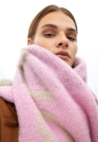 Écharpe rose et beige à motifs, en tissu doux. Présente une texture épaisse et confortable, drapée autour du cou, avec des fils visibles et des couches.