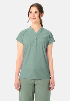 Femme portant une chemise boutonnée à manches courtes vert clair et un pantalon, debout et regardant vers le haut à sa gauche avec un léger sourire.