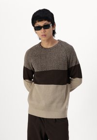 Maglione a maglia con una trama a coste, in strisce orizzontali di colore sabbia, marrone e marrone scuro. Collo a giro e fit rilassato. Abbinato a pantaloni scuri.