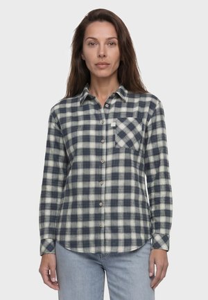 Frau mit langen braunen Haaren, die ein marineblau-weiß kariertes Hemd und hellblaue Jeans trägt und vor einem schlichten Hintergrund steht.