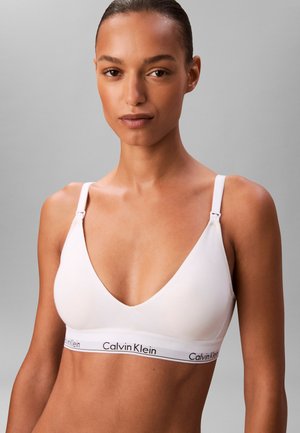 Kobieta prezentująca biały bralet Calvin Klein z regulowanymi ramiączkami na jednolitym szarym tle, zwrócona przodem.