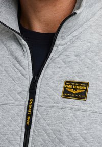 Grijze fleece zip-up jack met een gequilte patroon, zwarte rits en een geel- en zwartkleurig logo op de borst met het etiket "PME LEGEND."