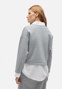 Sudadera de forro polar gris con hombros caídos, diseño cropped. Camisa blanca superpuesta visible en el dobladillo y los puños. Textura suave, costuras mínimas.