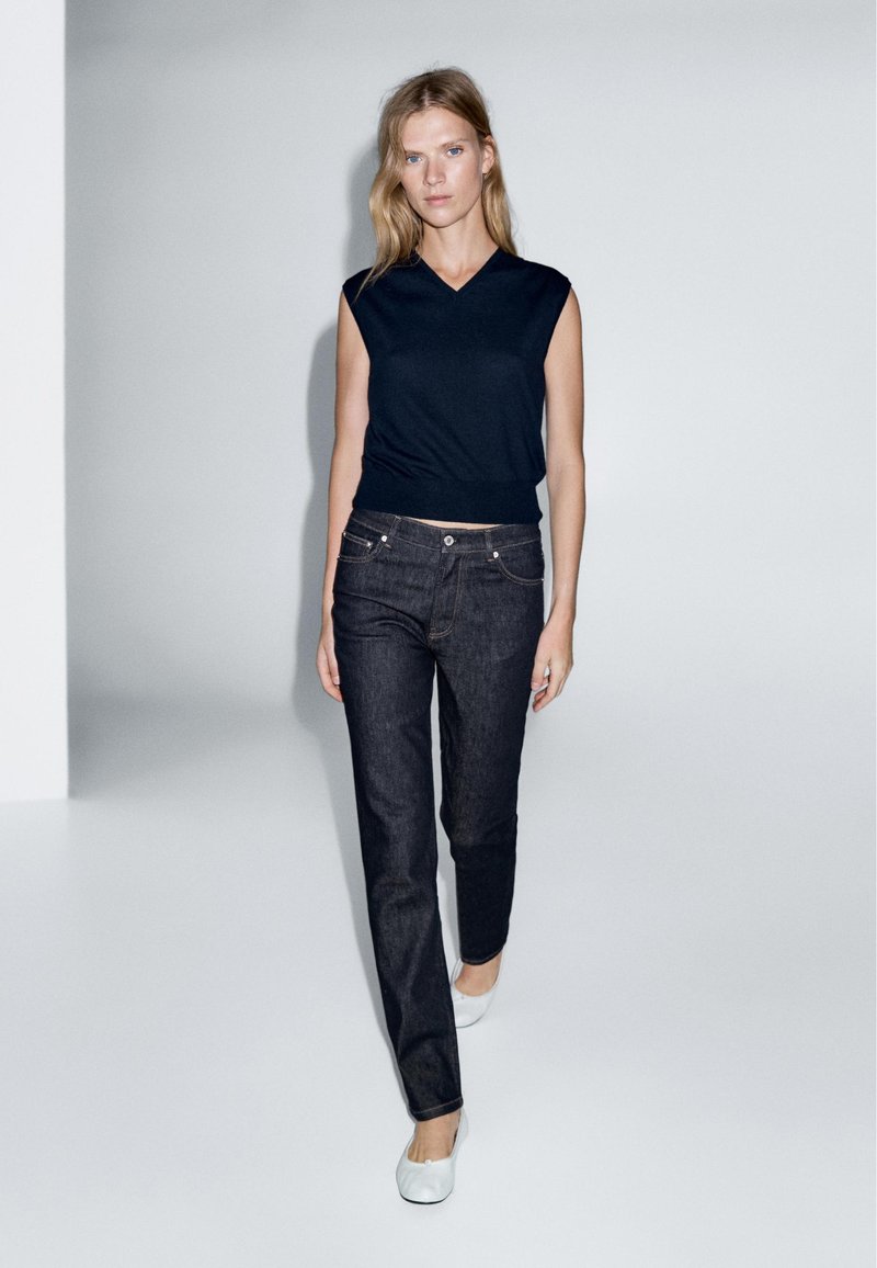 Top senza maniche blu navy, aderente in vita, abbinato a jeans dritti blu scuro. Il modello indossa delle ballerine bianche, in un ambiente minimalista.
