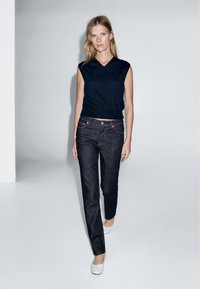 Granatowy bezrękawnik, dopasowany w talii, w zestawieniu z ciemnoniebieskimi prostymi dżinsami. Modelka nosi białe baleriny, stojąc w minimalistycznej scenerii.