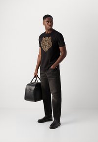 Black T-shirt with gold tiger graphic, dark jeans, and black leather bag. Model wears black chelsea boots, standing in a neutral backdrop.