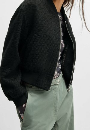 Černá bomber bunda se zipem a texturou nošená přes vzorované černé tričko, kombinovaná se světle zelenými kalhotami s vysokým pasem a páskem.
