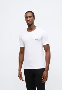 Baltas trumpomis rankovėmis medvilninis marškinėlis su apvalia iškirpte ir mažuoju raudonu "HUGO" logotipu ant krūtinės. Standartinis siluetas.