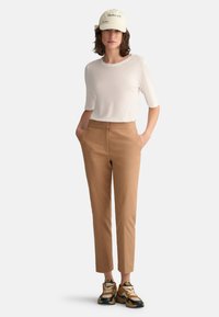White short-sleeve top, tan tailored pants, and black/gold sneakers, complemented by a light beige cap with "GANT" logo, standing with hands in pockets.