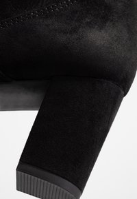 Lähiotoksessa musta mokkanahkaisen lohko korkokengän rakenne ja kumipohjan yksityiskohta valkoista taustaa vasten.