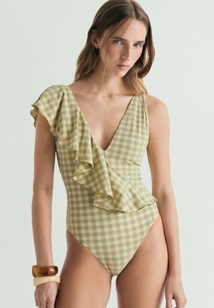 Mujer con bañador de un solo hombro a cuadros en verde y beige con volantes y dos pulseras gruesas en la muñeca izquierda, fondo neutro.