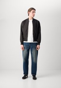 Schwarze Bomberjacke mit Reißverschluss und gerippten Bündchen, getragen über einem weißen T-Shirt. Kombiniert mit ausgewaschenen blauen Jeans und schwarzen Sneakers.
