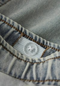 Jeans tkanina svetlo modre barve z vidno teksturo, na kateri je jasen gumb z rastlinskim vzorcem in rjavim šivom ob robu.