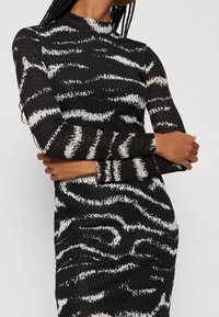 Femme portant une robe longue à manches longues texturée noir et blanc avec des rayures abstraites ressemblant à celles d'un tigre, les bras croisés à la taille.