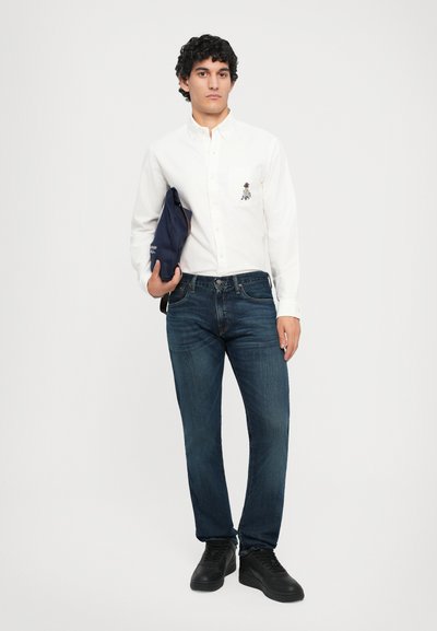 Cămașă albă cu butoni, cu buzunar și design brodat, purtată cu blugi negri și adidași negri. Modelul ține o geantă bleumarin.