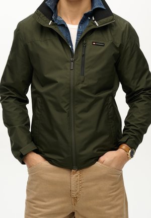 Homme portant une veste zippée vert olive sur une chemise bleue et un tee-shirt blanc, avec un pantalon beige et une montre-bracelet en cuir marron. Mains dans les poches.