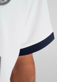 Arm with brown skin wearing a white short-sleeve shirt with a navy blue cuff, against a plain light gray background.
