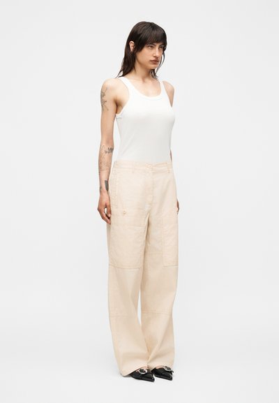 Γυναίκα με σκούρα μαλλιά και τατουάζ που φοράει λευκό αμάνικο τοπ, μπεζ παντελόνι με φαρδιά πόδια και τσέπες και μαύρα μυτερά παπούτσια με ασημένια αγκράφα.