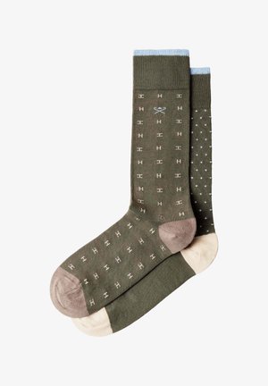 Par de calcetines verde oliva con puños azules, talones y dedos beige; un calcetín tiene un pequeño patrón de "H", el otro está salpicado de pequeñas cruces.