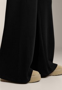 Crne široke hlače izrađene od mekanog materijala, s kontrastnim platformskim cipelama u boji kože s teksturiranim đonom i vidljivim detaljima šavova.