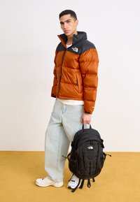 Padded orange and black jacket with a high collar, paired with light blue jeans and a black backpack. White sneakers complete the outfit.