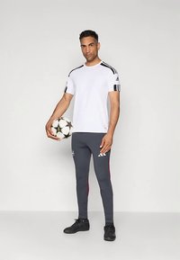 Baltas sportinis marškinėlis su juodomis juostelėmis ant pečių, derinamas su pilkomis tamprėmis su raudonomis šoninėmis detalėmis. Modelis laiko futbolo kamuolį.