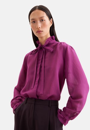 Donna con lunghi capelli scuri che indossa una blusa viola trasparente a maniche lunghe con fiocco al collo e pantaloni scuri a vita alta.