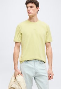 Νεαρός άνδρας που φοράει ανοιχτό κίτρινο πουκάμισο με κοντά μανίκια και ανοιχτό μπλε τζιν, κρατώντας ένα μπεζ σακάκι στο αριστερό του χέρι μπροστά σε απλό φόντο.