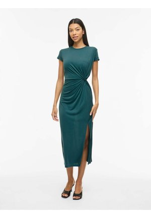 Kobieta ubrana w turkusową sukienkę do kolan z krótkimi rękawami, zębem zawiązanym z przodu z boku, rozcięciem do uda i czarnymi sandałami z odkrytymi palcami, stojąca na białym tle.