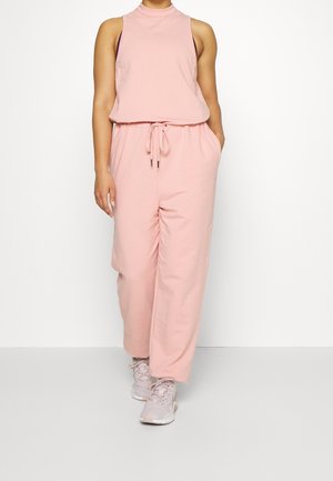 Person trägt einen ärmellosen, hellrosa Jumpsuit mit Taillen-Kordelzug und hellrosa Sneakers und steht vor einem schlichten weißen Hintergrund.
