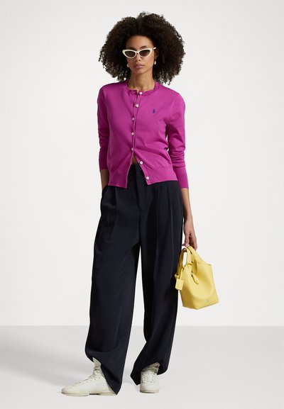 Cardigan fucsia realizzato in morbido tessuto a maglia, con scollatura rotonda e chiusura a bottoni. Abbinato a pantaloni larghi neri e una borsa gialla.