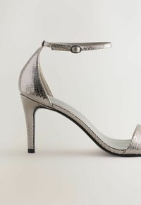 Metalické strieborné sandále na vysokom opätku s textúrovaným vzorom hadiej kože, otvorenou špičkou, pásikom okolo členka so sponou a elegantným zakriveným siluetou.