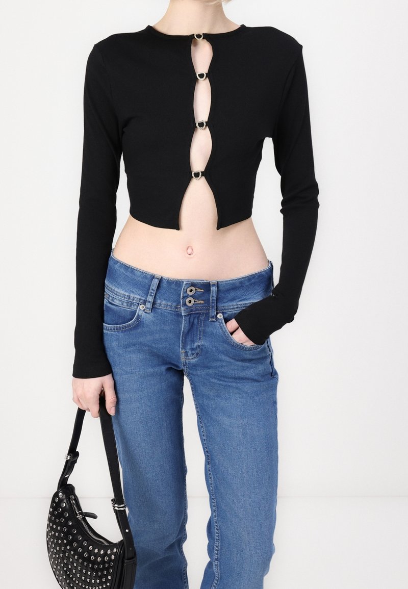 Femme portant un crop top noir à manches longues avec quatre découpes et anneaux, jean bleu taille basse, tenant un sac à main noir clouté.