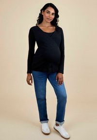 Schwarzes geripptes Wickeltop über einem schwarzen Tanktop, kombiniert mit blauen Jeans und weißen Sneakers. Das Top hat lange Ärmel und eine figurbetonte Passform.