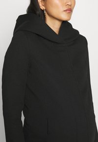 Manteau noir avec une capuche à col haut, fabriqué en tissu doux. Présente un design épuré et des coutures minimales, créant un aspect élégant.