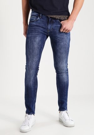 Homme portant un jean slim bleu foncé, des baskets blanches et une chemise noire, ajustant la taille du pantalon d'une main.