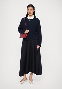 Pull en maille bleu marine avec un ourlet côtelé, superposé à une chemise blanche à col. Jupe noire fluide, accompagnée de ballerines à talons beiges et d'un sac à main rouge.