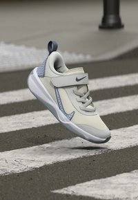 Hellgrauer Sportschuh mit einem Netz-Obermaterial, gepolstertem Schaft, Klettverschluss und dezenten blauen Akzenten, ausgestattet mit einer strukturierten Sohle für besseren Halt.