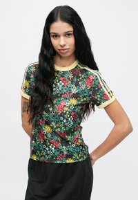 Γυναίκα με μακριά μαύρα μαλλιά που φοράει μαύρο πουκάμισο με φλοράλ σχέδιο και κίτρινο περιθώριο στα μανίκια, καθώς και μαύρο παντελόνι, στέκεται με το ένα χέρι στη μέση.