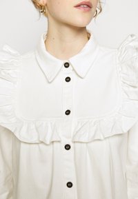 Hvid bomuldsskjorte med flæse langs kraven og knapstolpen, med sorte knapper. Stoffet har en subtil tekstur og et struktureret design.