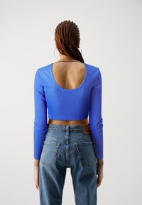 Un crop top bleu à côtes avec des manches longues, doté d'un grand dos ouvert. Assorti à un jean en denim taille haute, mettant en valeur des poches arrière emblématiques.