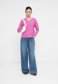 Sweater de malha canelada fúcsia com decote em V, combinado com jeans de perna larga azuis. Modelo segura uma pequena bolsa branca. Fundo simples.