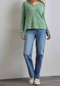 Pull vert duveteux avec motif géométrique beige, associé à un jean bleu clair et des baskets beige avec des accents blancs. Design à col en V.